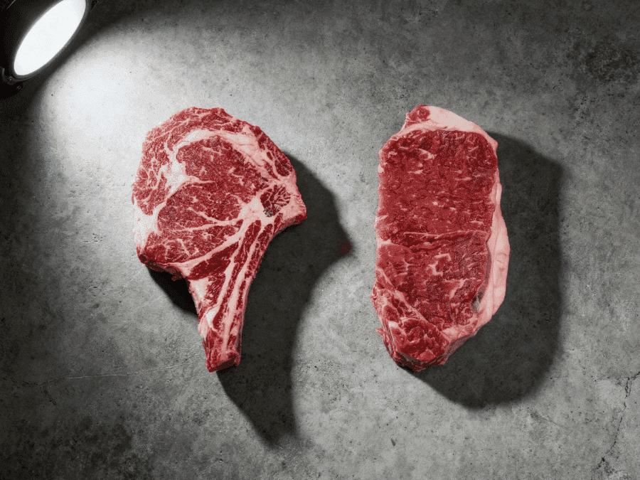 Ribeye vs strip steaks side by side on concrete surface showing marbling difference — Boucherie Beirut