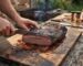 Person slices a smoked beef brisket on a wooden cutting board outdoors, smoke rising as juices pool nearby.